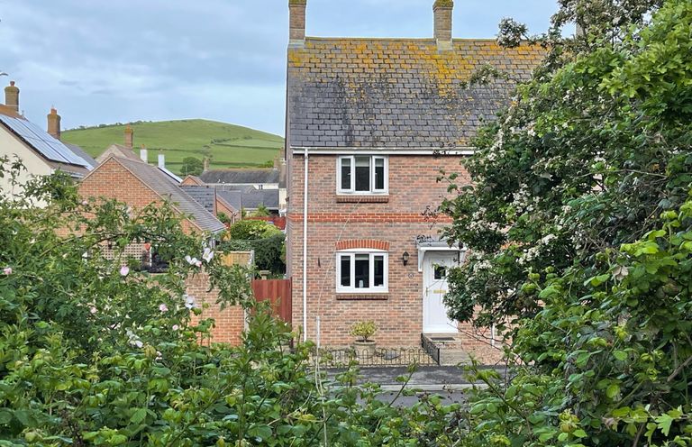 A brick house with white windows and door surrounded by green bushes and trees with a hill in the background at The Beach House in West Bay