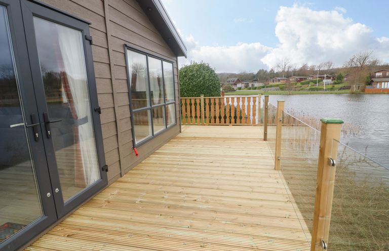 A wooden deck attached to a house overlooking a lake with glass and wood railings at Elim in South Lakeland Leisure Village near Tewitfield