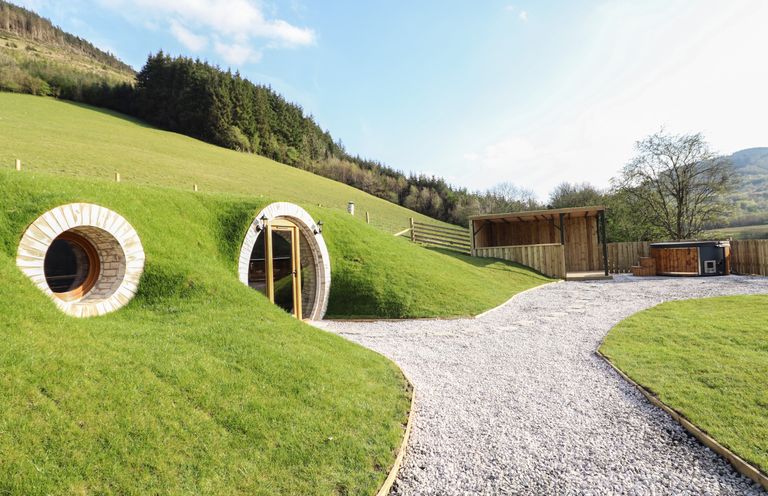 A hillside home with round windows and door next to a gravel path and wooden shelter at Nant y Garnedd Mallwyd near Dinas Mawddwy