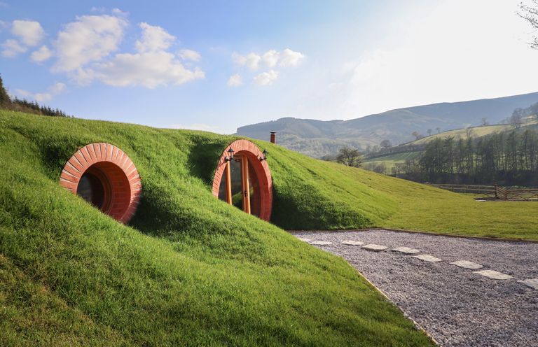 A grassy earth house with round doors and windows in a rural setting with hills and a gravel path at Nant yr Esgair Mallwyd near Dinas Mawddwy