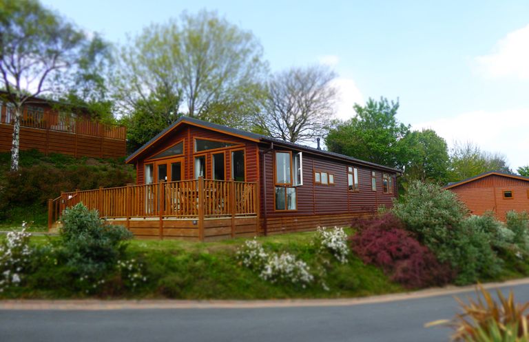 A wooden cabin with a fenced deck surrounded by bushes and trees at 23 Bossiney Bay in Tintagel