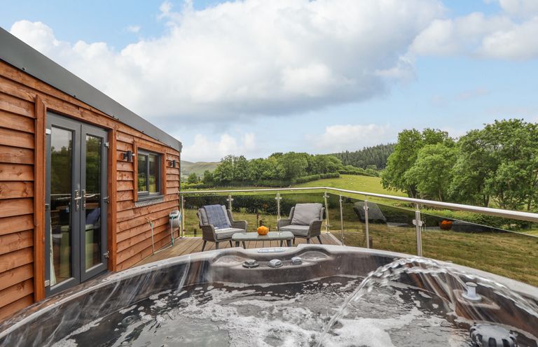 An outdoor deck with a hot tub chairs and a table overlooking green fields at The Cabin at Evelyns Retreat in Glyndyfrdwy
