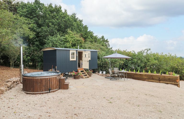 An outdoor area with a wooden hot tub a small black shepherds hut with steps a table with chairs and an umbrella at High Grounds Shepherd's Hut Boylestone near Ashbourne
