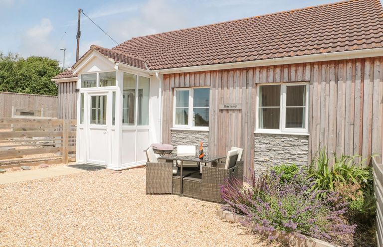 An outdoor seating area with a table and chairs in front of a wooden house with a tiled roof and a small garden at Garland in Nottington near Weymouth