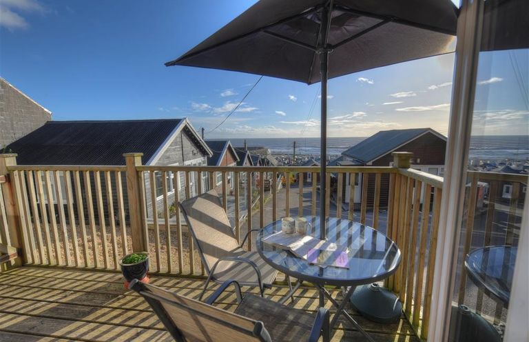 A wooden balcony with a table and chairs under an umbrella overlooking houses and the sea at St Anton in Lyme Regis