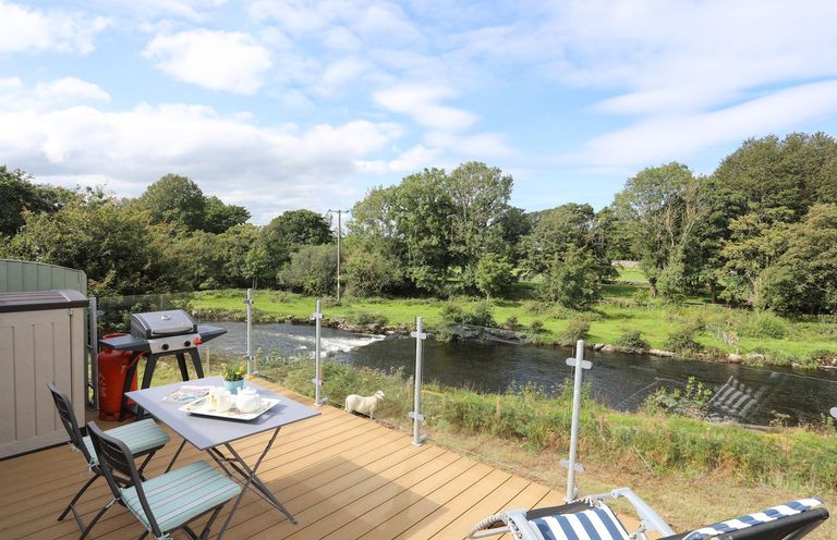 An outdoor deck with a table and chairs a barbecue and a striped lounge chair overlooking a river and trees at Llety'r Bugail 1 in Llanrug