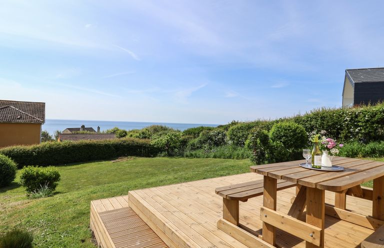 A wooden picnic table on a deck overlooking a grassy garden with bushes and houses near the sea at Tillicum in Charmouth
