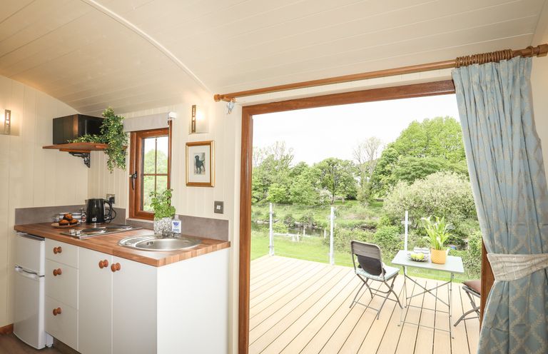 A small kitchen with a sink and countertop next to a glass door leading to a wooden deck with a table and chairs overlooking trees at Llety'r Bugail 2 in Llanrug