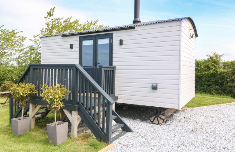 A small white cabin with black double doors and a chimney on a gravel area with stairs and plants at Cwt Y Rhos in Valley
