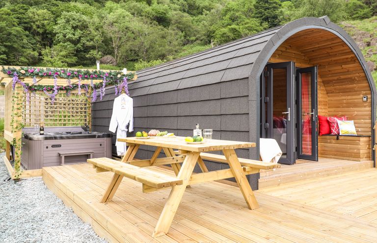 A wooden patio with a picnic table hot tub and arched cabin in a forested area at Hartsop Magic - Crossgate Luxury Glamping Hartsop near Glenridding