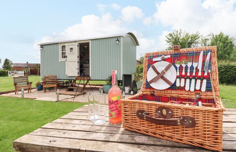 A picnic basket with cutlery and plates on a wooden table outside a small green cabin with chairs on a lawn at Honeybee Lodge in Stixwould near Woodhall Spa