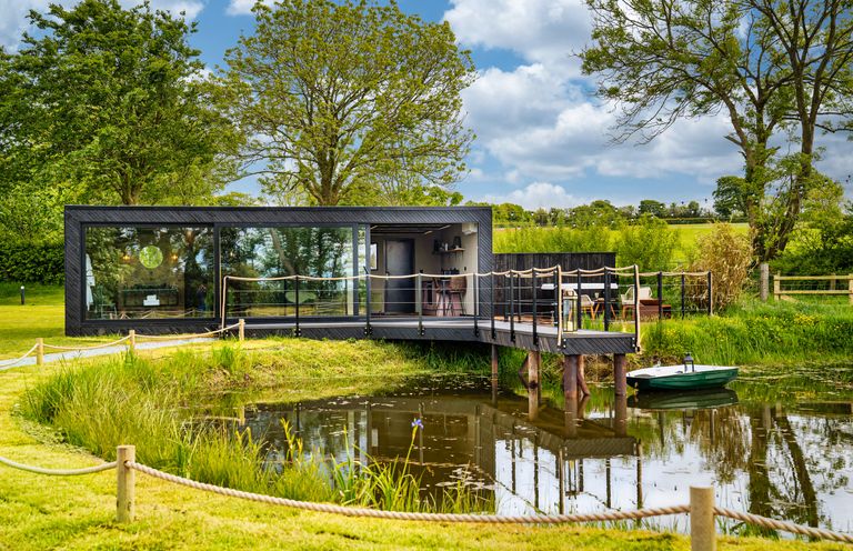 A modern black cabin with large glass windows beside a pond with a small boat and surrounded by green trees and grass at Rusty Boathouse in Llanarth