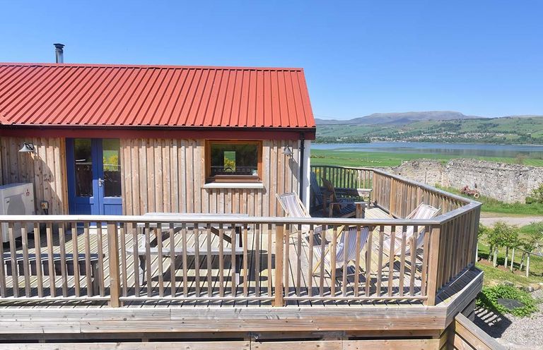 A wooden deck with chairs and a table outside a house with a red roof overlooking a body of water at Skylarks Ferintosh near to Dingwall