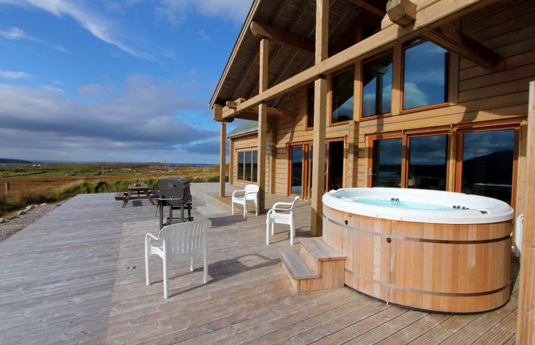 An outdoor wooden deck with a hot tub white plastic chairs a BBQ grill and a picnic table at Eagle Lodge in Uig near Stornoway