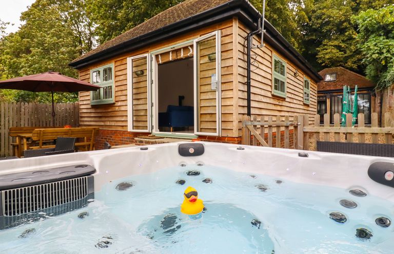 An outdoor hot tub with a rubber duck in front of a wooden cabin with open doors and outdoor seating with an umbrella at Dreamwood Lodge in Keston near Leaves Green