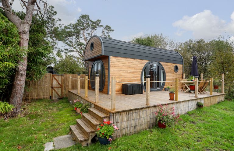 A wooden cabin with a deck surrounded by trees and grass at The Hideaway Pod in Rhoslan near Criccieth