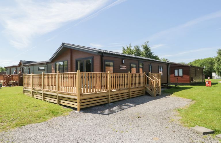 A wooden lodge with a fenced porch and gravel driveway on green grass under a blue sky at Parkers Place in Carnforth