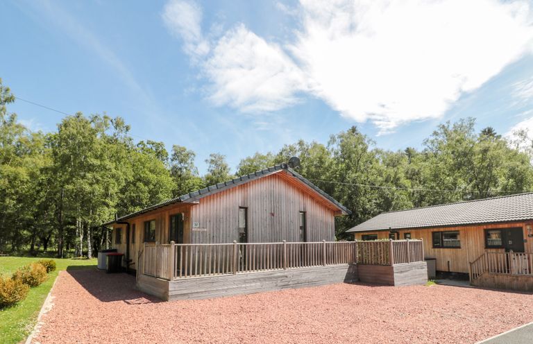 A wooden cabin with a fenced porch surrounded by trees and gravel in front at Lodge 32 Otterburn