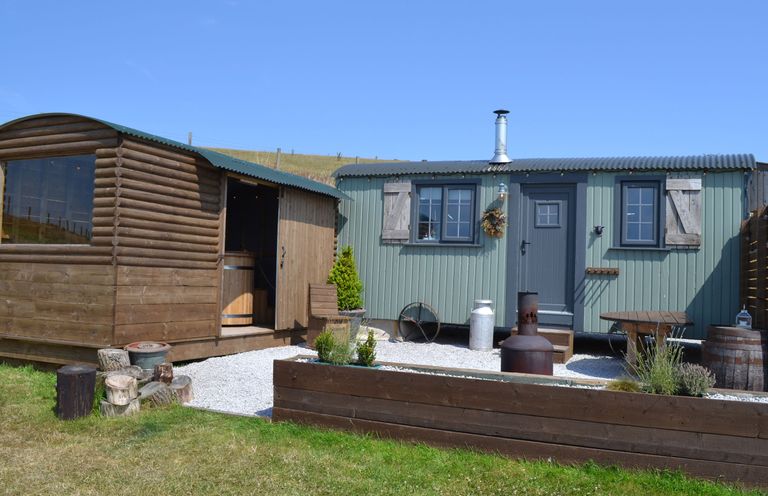 An outdoor area with two small buildings a wooden cabin and a green metal hut with a chimney and garden beds at Heather Hut at Copy House Hideaway in Earby
