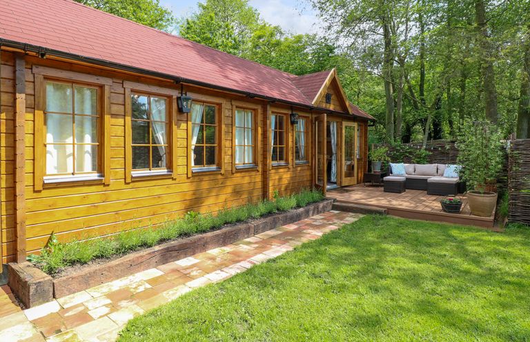 A wooden cabin with multiple windows and a red roof next to a patio with outdoor furniture and a garden with grass and plants at Willow Lodge in Potter Heigham
