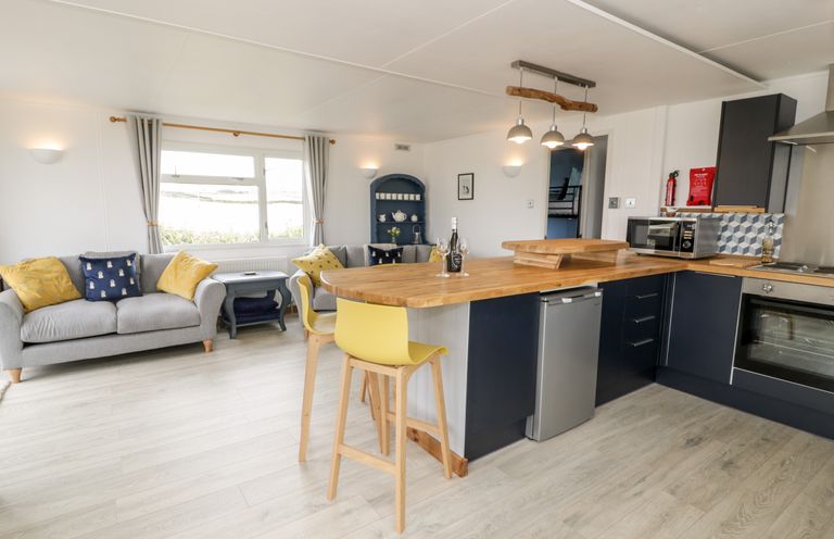 An open plan kitchen and living area with wooden countertops yellow chairs and grey sofas at Porthwen Lodge in Bull Bay near Cemaes Bay