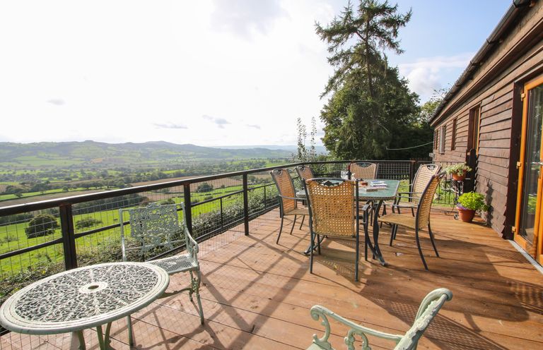 A wooden deck with metal and wicker chairs and tables overlooking green fields and hills at Dragonfly Lodge in Worthen near Minsterley