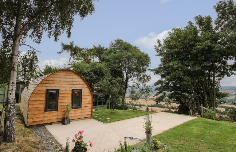 A small wooden pod with two windows on a concrete pad surrounded by grass and trees at The Pod in Worthen near Minsterley