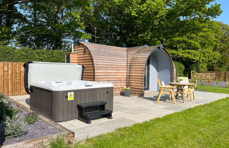 A wooden pod structure with a hot tub patio and table with two chairs on grass at Yr Onnen in Bontuchel near Ruthin