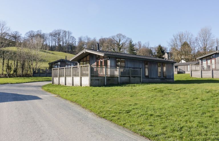 A wooden cabin with a fenced porch on a grassy plot near a gravel road at Rivendell Lodge in Roger Ground near Hawkshead