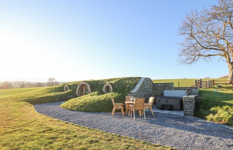 An outdoor grassy earth shelter with round windows a wooden table with four chairs and a hot tub at Tan y Wawr- Onnen in Llanfair Caereinion