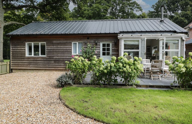 A single story wooden house with a gravel driveway and green plants in front at The Roost in Cheriton near Alresford