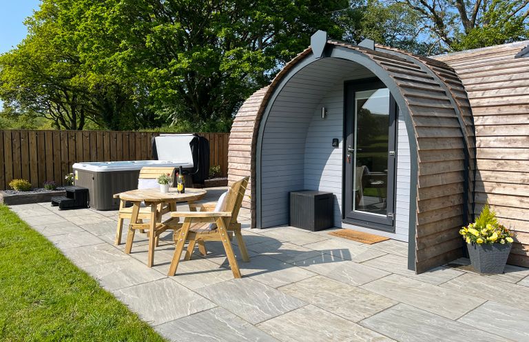 A wooden pod with a glass door on a stone patio with a hot tub wooden table and chairs and a flower pot at Y Dderwen in Bontuchel near Ruthin
