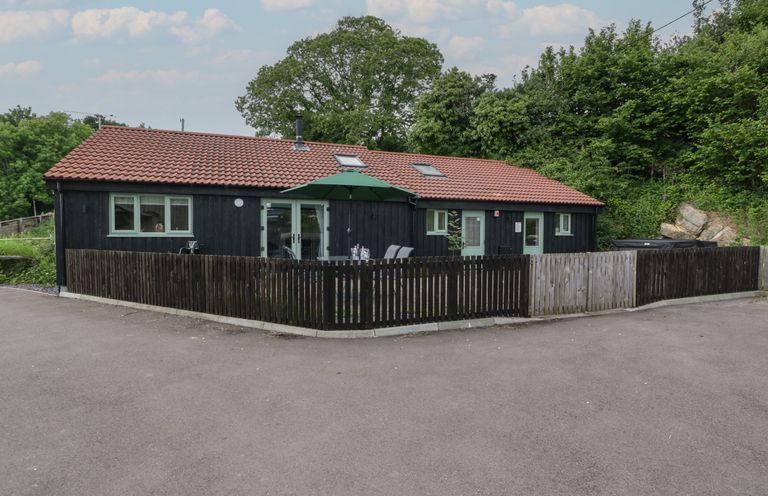 A single story building with a tiled roof and fenced patio with an umbrella and chairs surrounded by trees at Bottom Lodge in Penhow