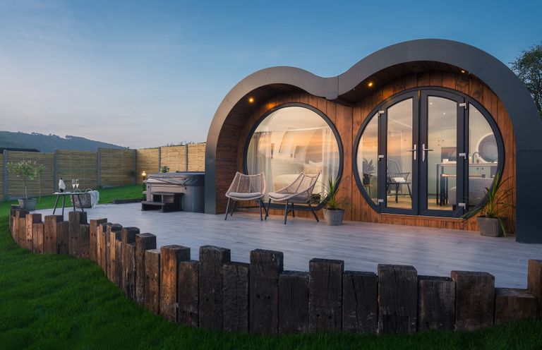 A modern wooden cabin with large round windows and a patio with chairs and a hot tub at Creuddyn in Llanfihangel-y-Creuddyn near Aberystwyth