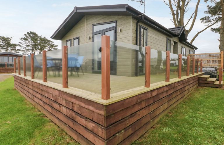 A raised wooden deck with glass railing around a small house on grass at Driftwood Lodge in Hasguard Cross near Broad Haven