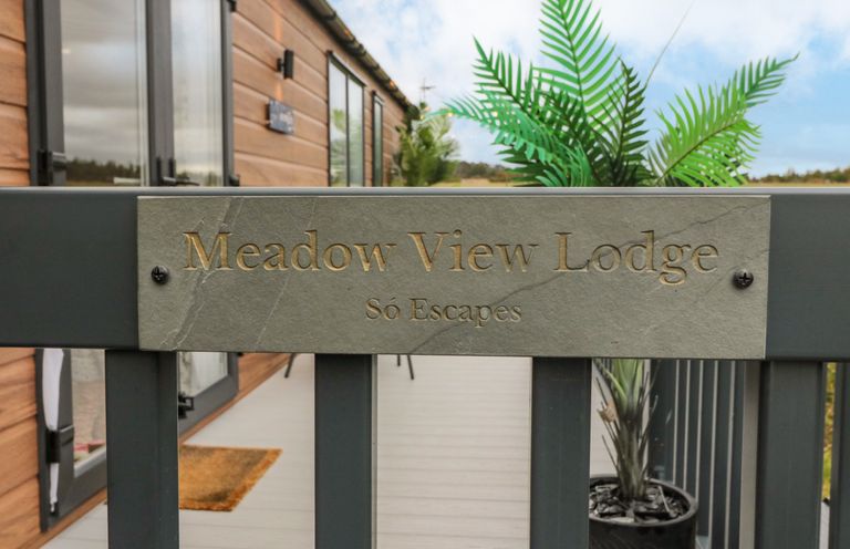 A gate with a Meadow View Lodge sign in front of a wooden lodge with glass doors and a potted plant on the porch at Plot 18 Meadow View in Felton
