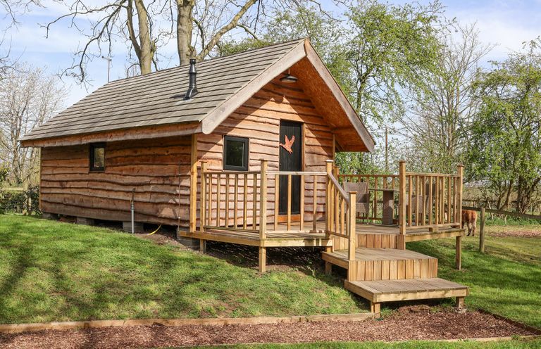 A wooden cabin with a small porch and stairs in a grassy area with trees at Duck Lodge in Bedale