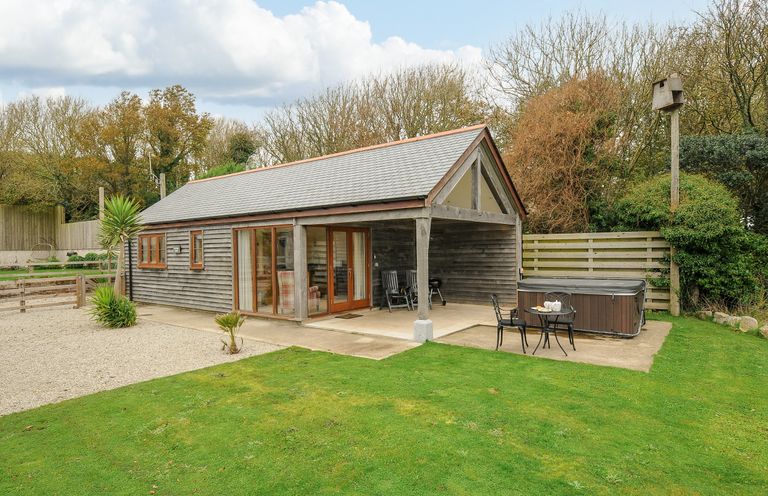 An outdoor view of a wooden cabin with a covered patio two chairs a small table with two chairs and a hot tub in the yard at Pol Glas Cabin in Churchtown near Mullion