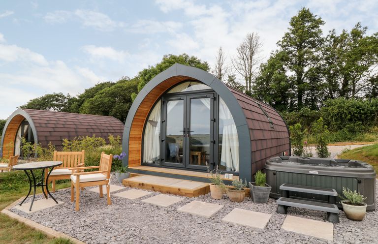 Two arched wooden cabins with glass doors a patio set and a hot tub surrounded by plants at Ash in Saltash