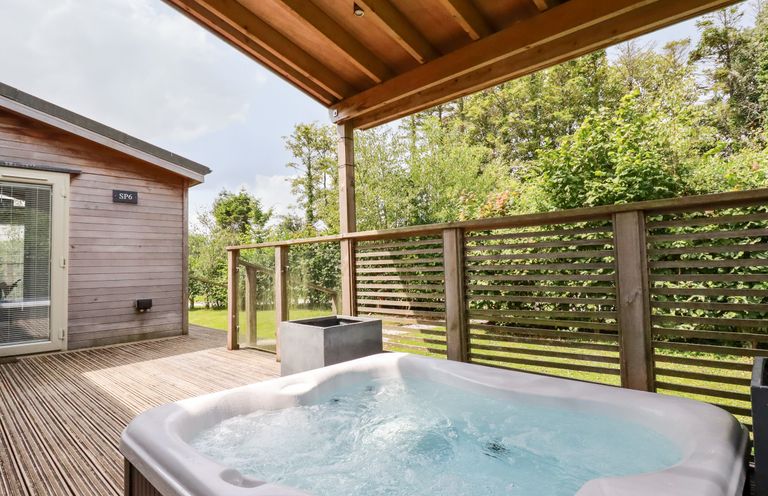 An outdoor wooden deck with a hot tub under a covered roof and wooden railing surrounded by greenery at 6 Southern Place in Dobwalls
