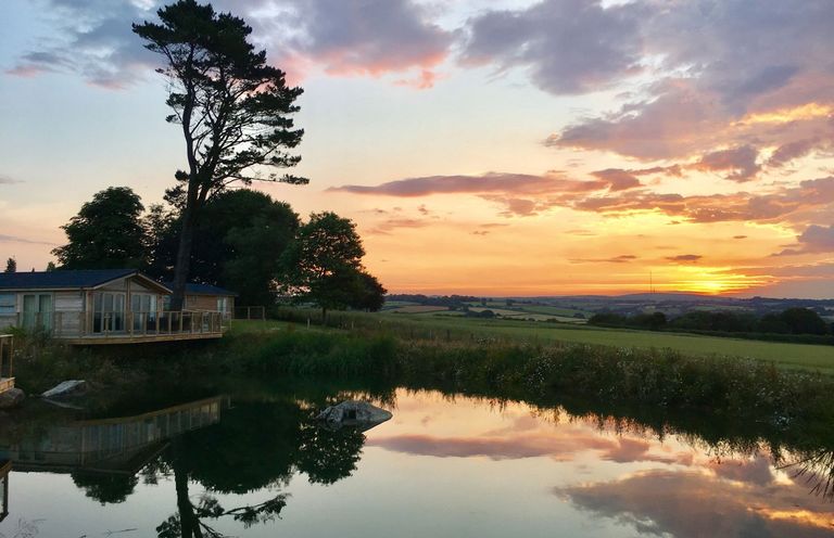 A pond reflecting trees and a cabin with a sunset over fields in the background at 12 Faraway Fields in Dobwalls