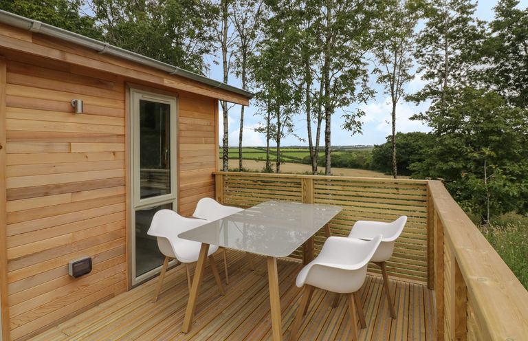 An outdoor wooden deck with a glass table and four white chairs overlooking trees and fields at 18 Meadow Retreat in Dobwalls