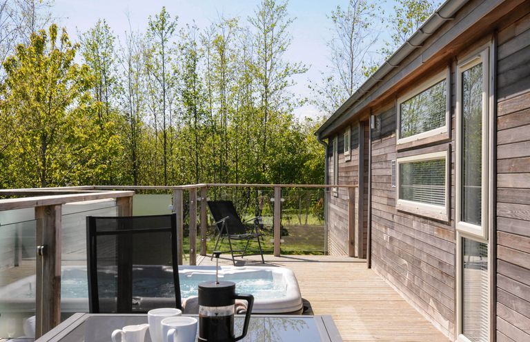 A wooden deck with a glass table and coffee set next to a hot tub and black chairs at 7 Meadow Retreat in Dobwalls