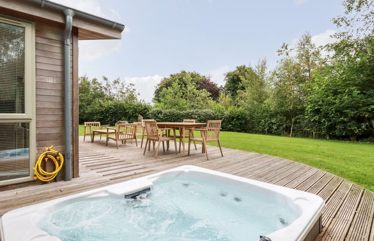 An outdoor area with a hot tub on a wooden deck and a wooden table with six chairs surrounded by grass and trees at 14 Horizon View in Dobwalls