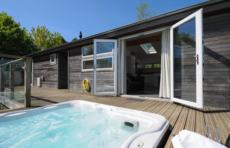 An outdoor hot tub on a wooden deck with glass railing and open double doors leading to a room at 5 Meadow Retreat in Dobwalls
