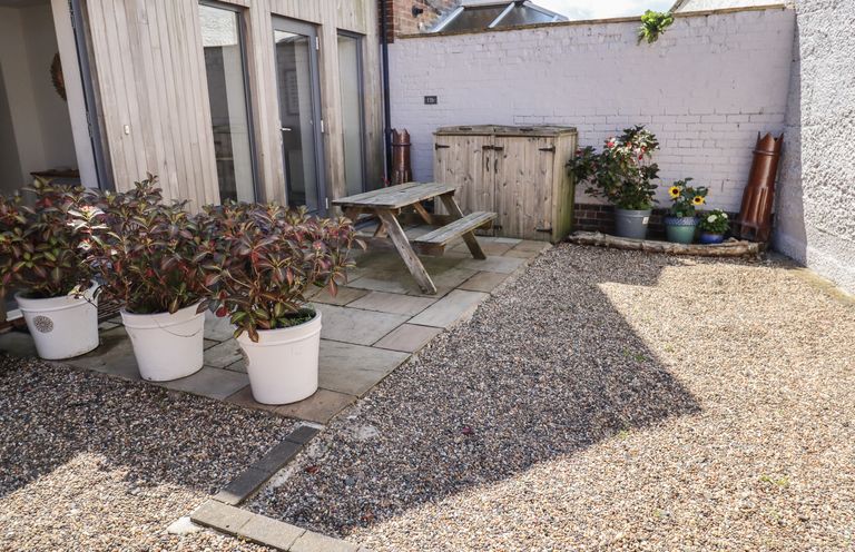 An outdoor patio area with potted plants a wooden picnic bench a storage box and gravel ground at Riverside Lodge in Alnmouth