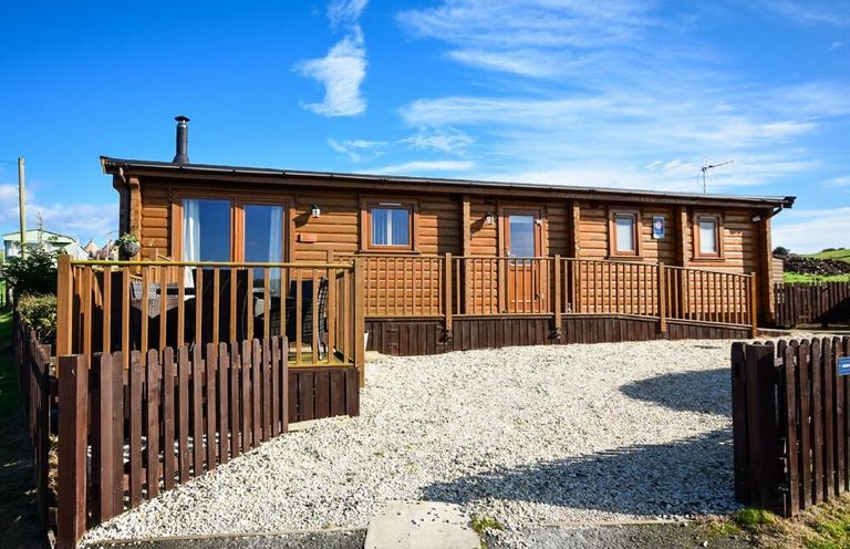 A wooden cabin with a fenced gravel driveway under a blue sky at Heather Cottages - Brown Owl in Bamburgh