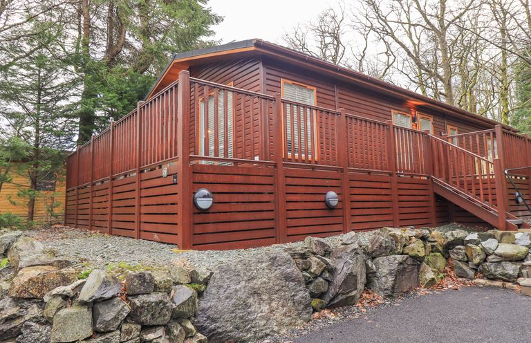 A wooden cabin with a raised deck and stairs surrounded by trees and a stone wall at 25 Thirlmere in Troutbeck Bridge