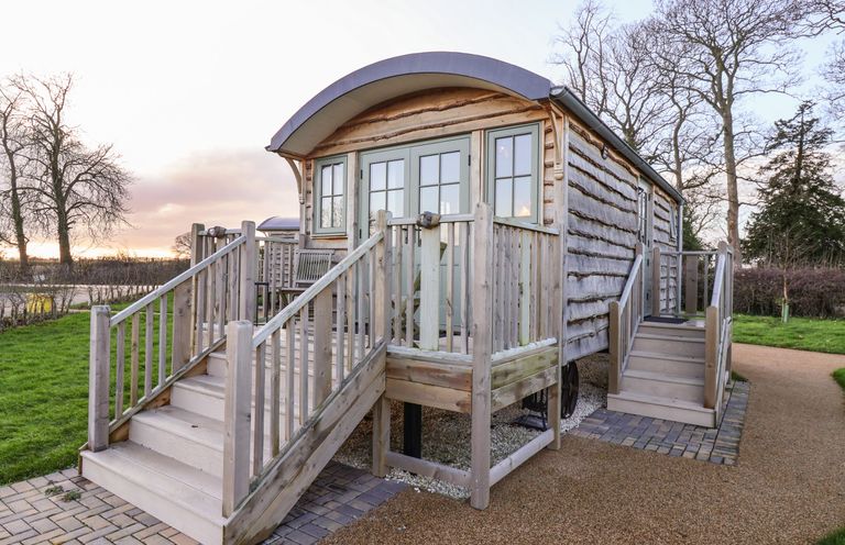 A wooden shepherd hut with two staircases and a small porch surrounded by trees and grass at The Storyteller in Bolton Percy near Tadcaster