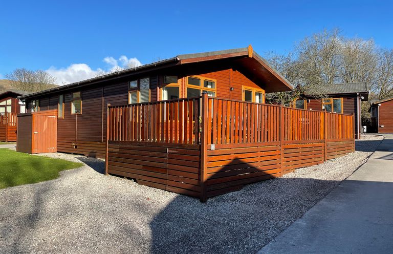 A wooden lodge with a fenced patio and gravel pathway at Highfield Lodge in Kirkstone 30 Limefitt Park near Troutbeck